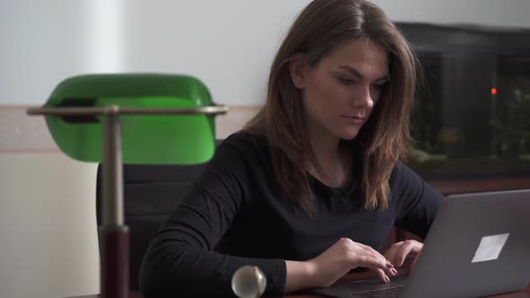 Young Woman with Long Hair Working at Office with Big Aquarium at Laptop Computer