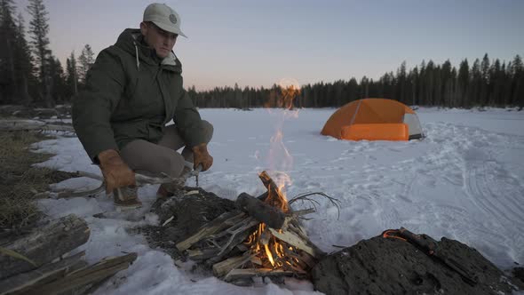 Man feeding a fire in the snow next to his tent. Tent in the snow next to a frozen lake.