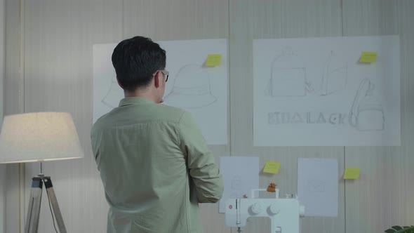 Back View Of Asian Man Designer Looking At The Bag Design Sketch Paper On The Wall