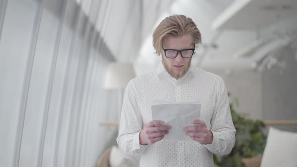 Portrait Successful Blond Man in Glasses Standing in a Light Comfortable Office Reading Papers