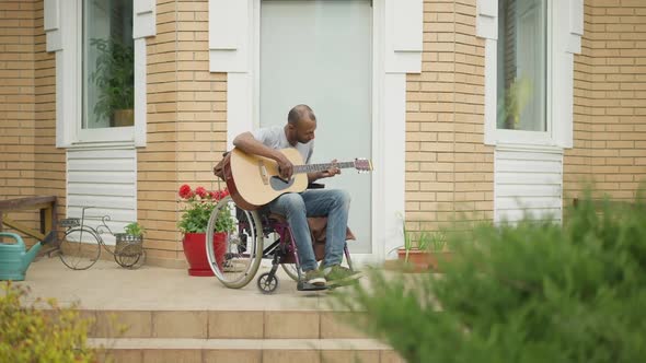 Wide Shot of Creative Disabled Man in Wheelchair Playing Guitar Singing on Backyard Porch