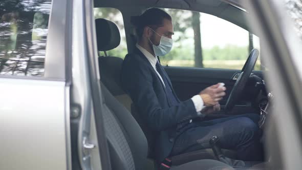 Side View Male Driver in Covid Face Mask Spraying Disinfectant on Steering Wheel Sitting on Driver's