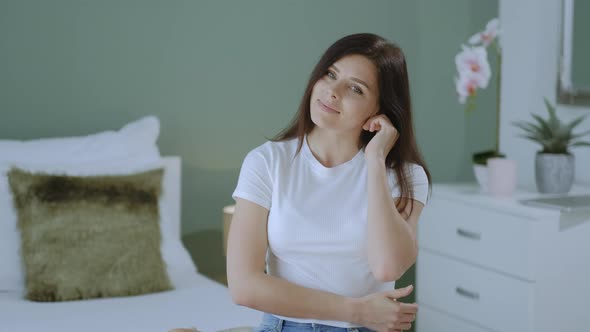 Pretty millennial brunette woman looking at the camera and smiling while sitting on bed at home