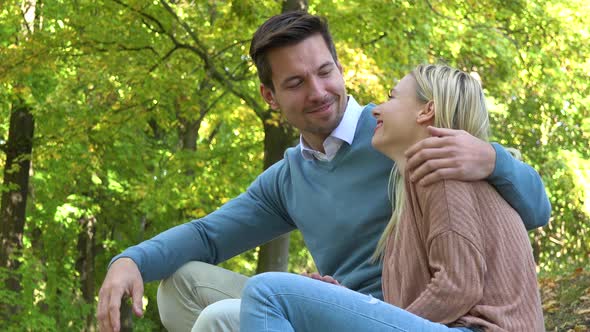 A Young Attractive Couple Sits in a Park on a Sunny Day and Waves at the Camera with a Smile