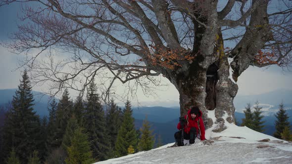 Man Tourist Rest Near a Big Tree in Winter Forest Snowy Background