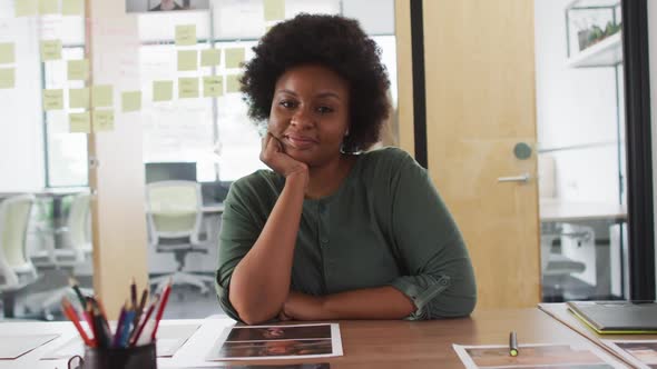 Portrait of african american businesswoman sitting at desk leaning on hand and smiling in office