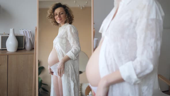 Happy Young Pregnant Woman Looking In Mirror On Her Belly. Motherhood, Pregnancy Concept.