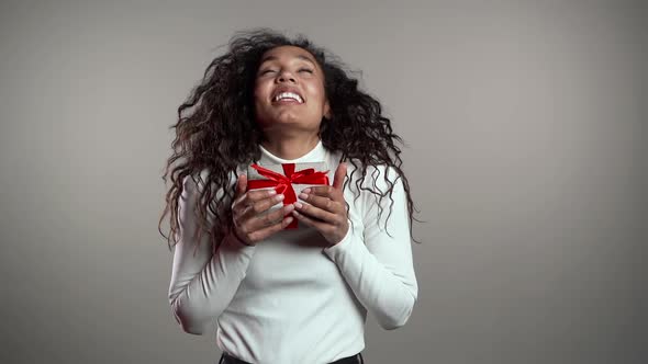 Surprised Mixed Race Woman Smiling, Holding Gift Box on Gray Studio Background
