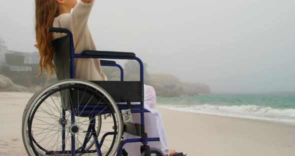 Side view of young Caucasian woman sitting with arms outstretched on wheelchair at beach 4k