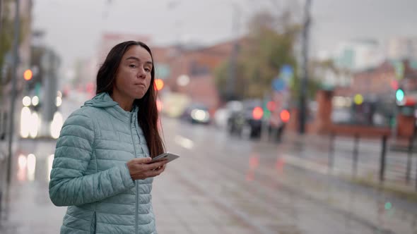 a Brunette in a Blue Jacket with Long Hair and with a Phone in Hand Stands on a Wet Street on a