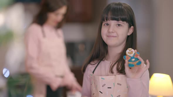 Cute Girl Boasting Gingerbread Cookie Standing in Kitchen with Blurred Woman at Background