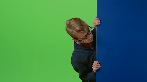 Teenager Boy in Glasses in a Sweater Peeks Out From Behind the Boards and Shows Like on a Green