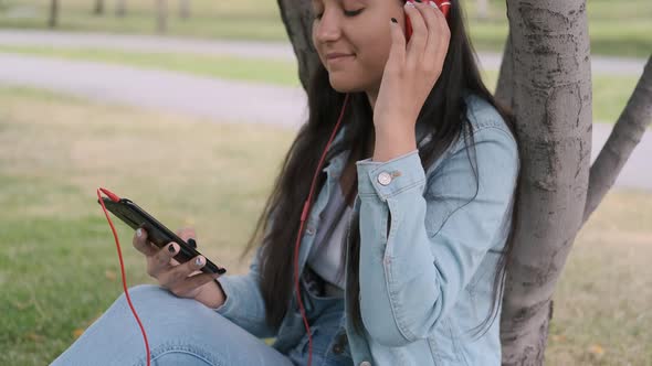 Girl Listening To Music in Headphones Sitting Near a Tree in the Park. Close-up