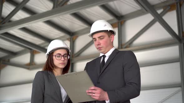 Young Man and Woman in Helmets with Documents at a Construction Site. Chiefs in Suits Discussing the