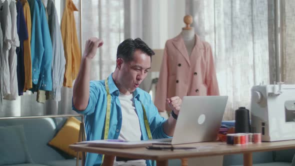 Happy Asian Male Designer Working On A Laptop And Celebrating While Designing Clothes In The Studio