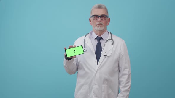 Adult man doctor in medical uniform shows the green screen of phone with app, standing in studio