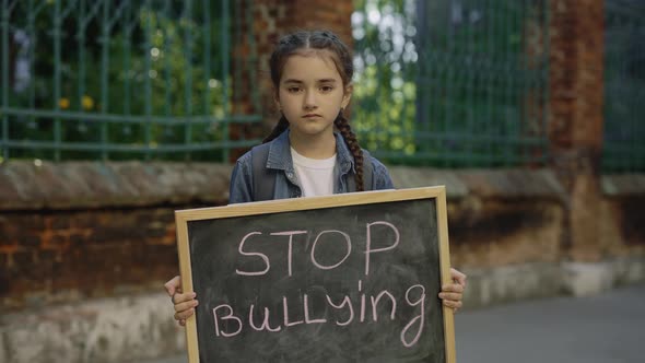 Portrait of Upset Little Girl Standing Holding Board with Text Message Stop Bullying on Street