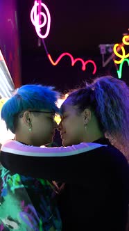 Cheerful lesbian girlfriends kissing against neon inscriptions