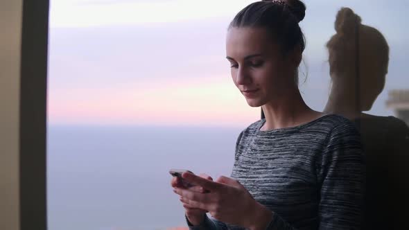 Closeup View of Beautiful Young Woman Sitting By the Open Window with a Smile Looking at Phone and