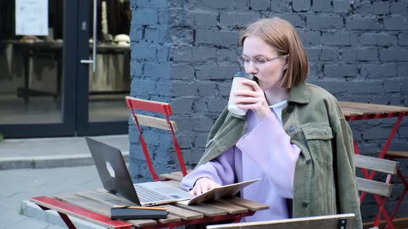 Thoughtful Young Businesswoman Typing on Laptop Outside in Street Cafe