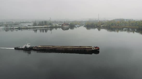 Barge sailing on wide river and transporting sand through bridge in smog