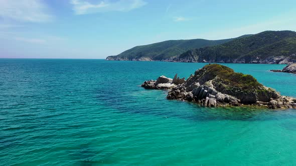 Small rocky island surrounded by crystal clear blue water with mountain and forest in the background