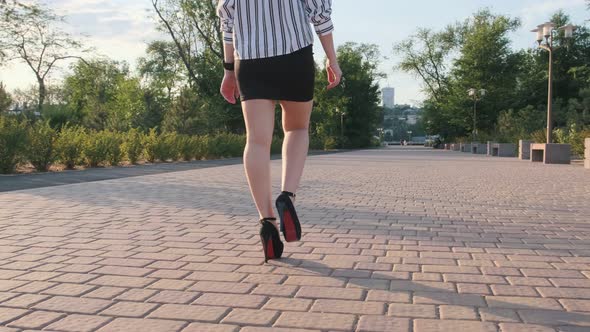 Slender female legs in black high-heeled shoes walk in the park on a sunny day. An elegant girl in a