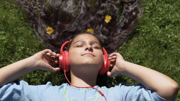 Attractive Girl with Long Black Hair Lies on the Grass in the Park and Listens to Music on