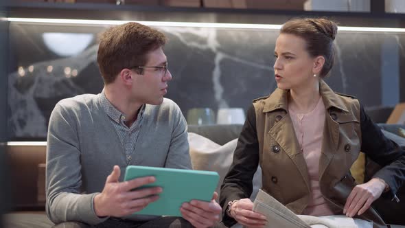 Front View Man with Tablet Talking to Woman Choosing Furniture Upholstery Samples in Showroom