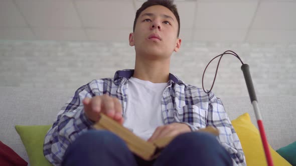 Blind Asian Young Man Reading a Book of Braille Text Sitting on the Couch in the Living Room Close