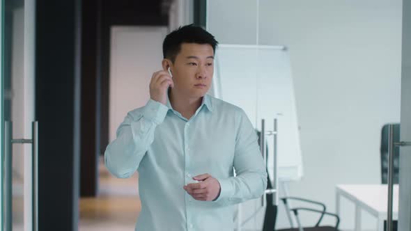 Positive Middle Aged Asian Man Manager Putting Earbuds Into Ears and Enjoying Great Music Standing