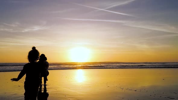 A child runs towards mom's open arms in slow-motion during a sunning sunset at the beach.