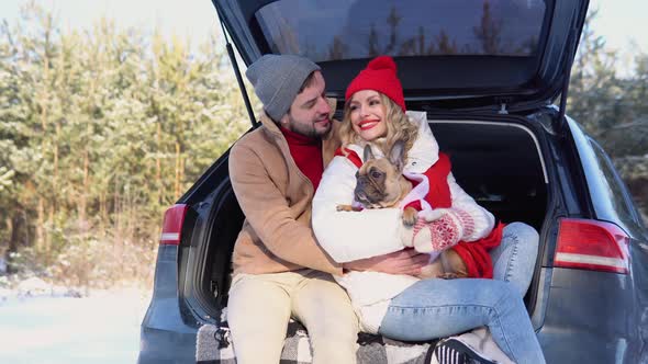 Young Couple a Man and a Woman with Dog are Sitting in the Trunk of a Car in a Winter and Hugging