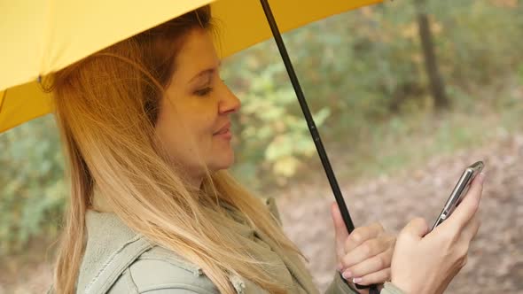 Slow motion blond  with yellow umbrella holding smart phone in the park 1080p HD footage - Blonde fe