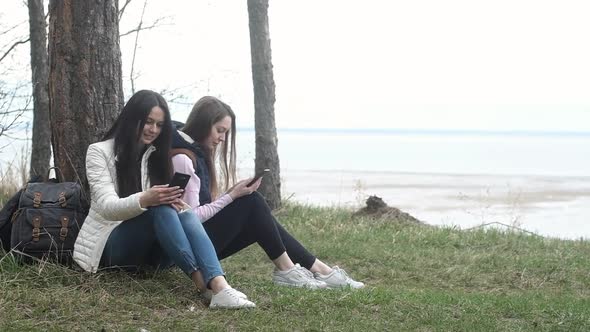 Two Caucasian female tourists sit cliff near tree and use their phones