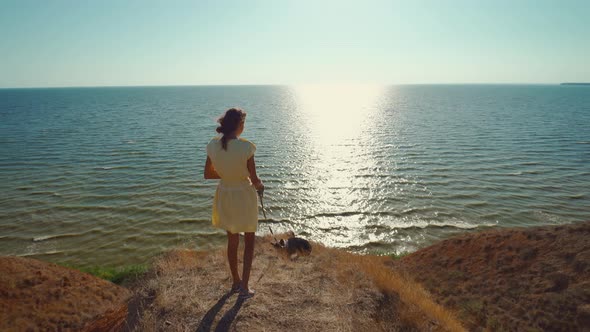 Rear View Beautiful Happy Girl in Yellow Dress with Cute Pet Corgi Dog Stands on Clay Hill with