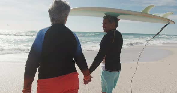 Back view of happy senior hispanic couple walking on beach with surfboard