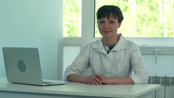 Female Doctor Working at Office Desk with Laptop and Smiling at Camera