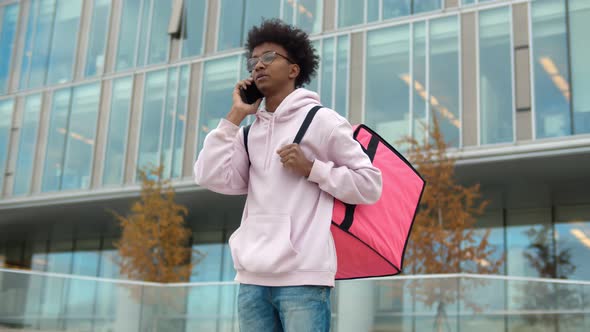 African Delivery Man with Backpack Talking on Phone To Client Standing Lost in Business District