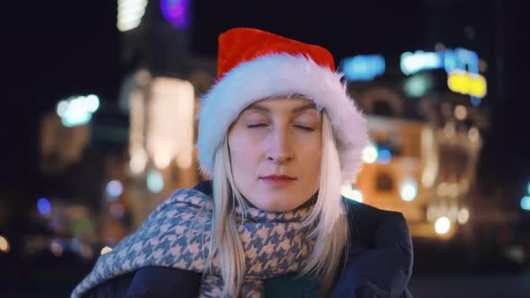 A young woman in a Santa hat looks into the camera . night city.