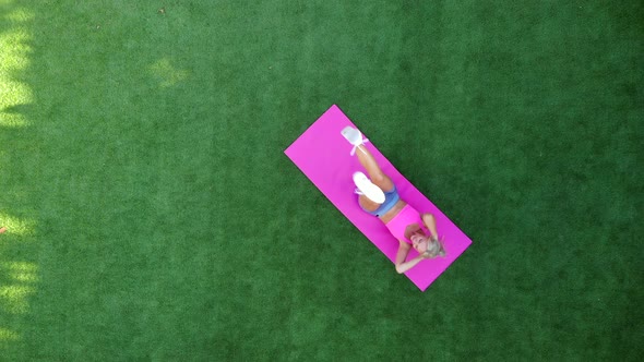 Top View Young Athletic Woman Trains Abdominal Muscles While Lying on Grass, Drone Shot