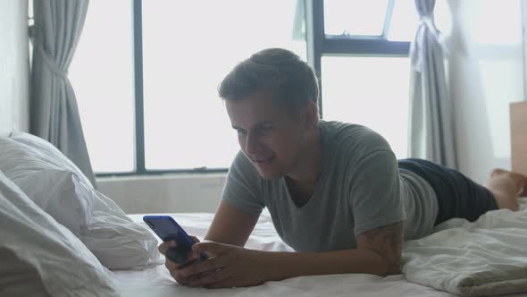 Young Handsome Man Lies in Bed By the Window Uses the Phone