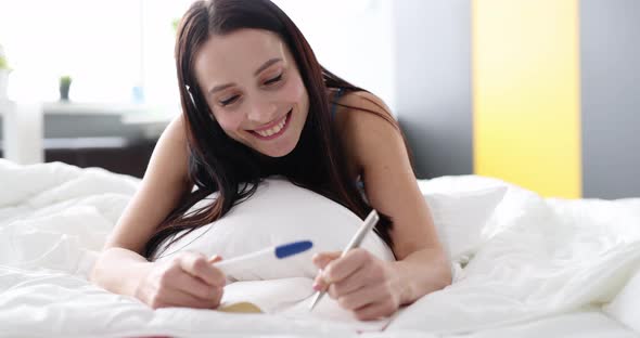 Happy Woman with Pregnancy Test in Bedroom Making Notes in Diary