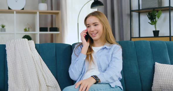 Lady with Blond Hair in Casual Clothes Sitting on Comfortable Sofa at Home and Talking on phone