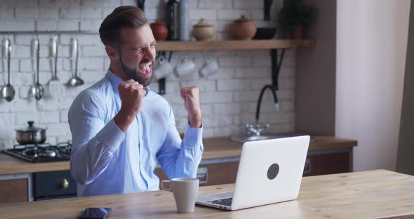 Young Business Man Sits at Table with a Laptop Working in Home Office Kitchen Background