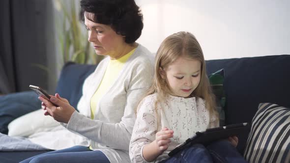 Senior Woman Is Sitting on Sofa and Looking at Smartphone While Young Girl Is Watching Something
