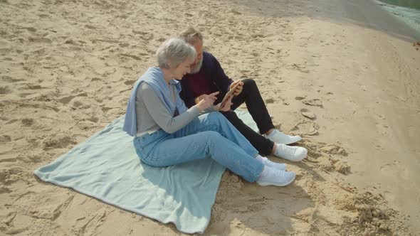 Aged Couple Sitting on Sand Watching Video or Photos Via Tablet