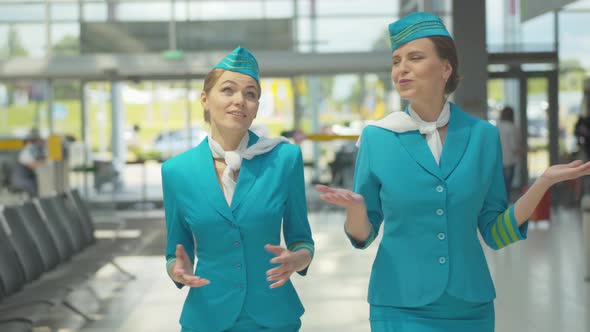 Middle Shot of Two Joyful Caucasian Women in Stewardess Uniform Walking for Departure in Airport