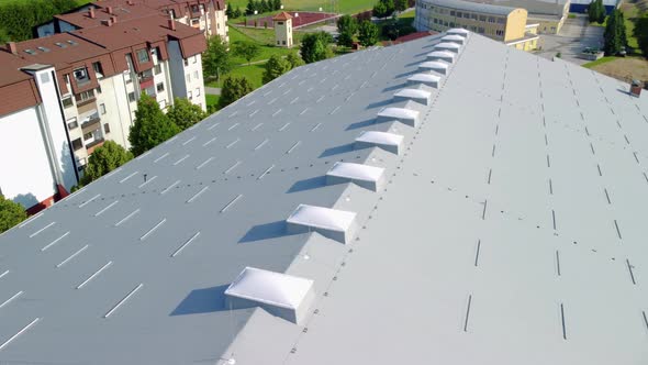 Flying over big sport hall covered with grey insulation panels and urbane blocks beside it. Aerial 4