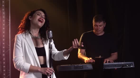 A Stylish Girl Vocalist Sings on Stage in a Vintage Microphone. In the Background a Man Playing